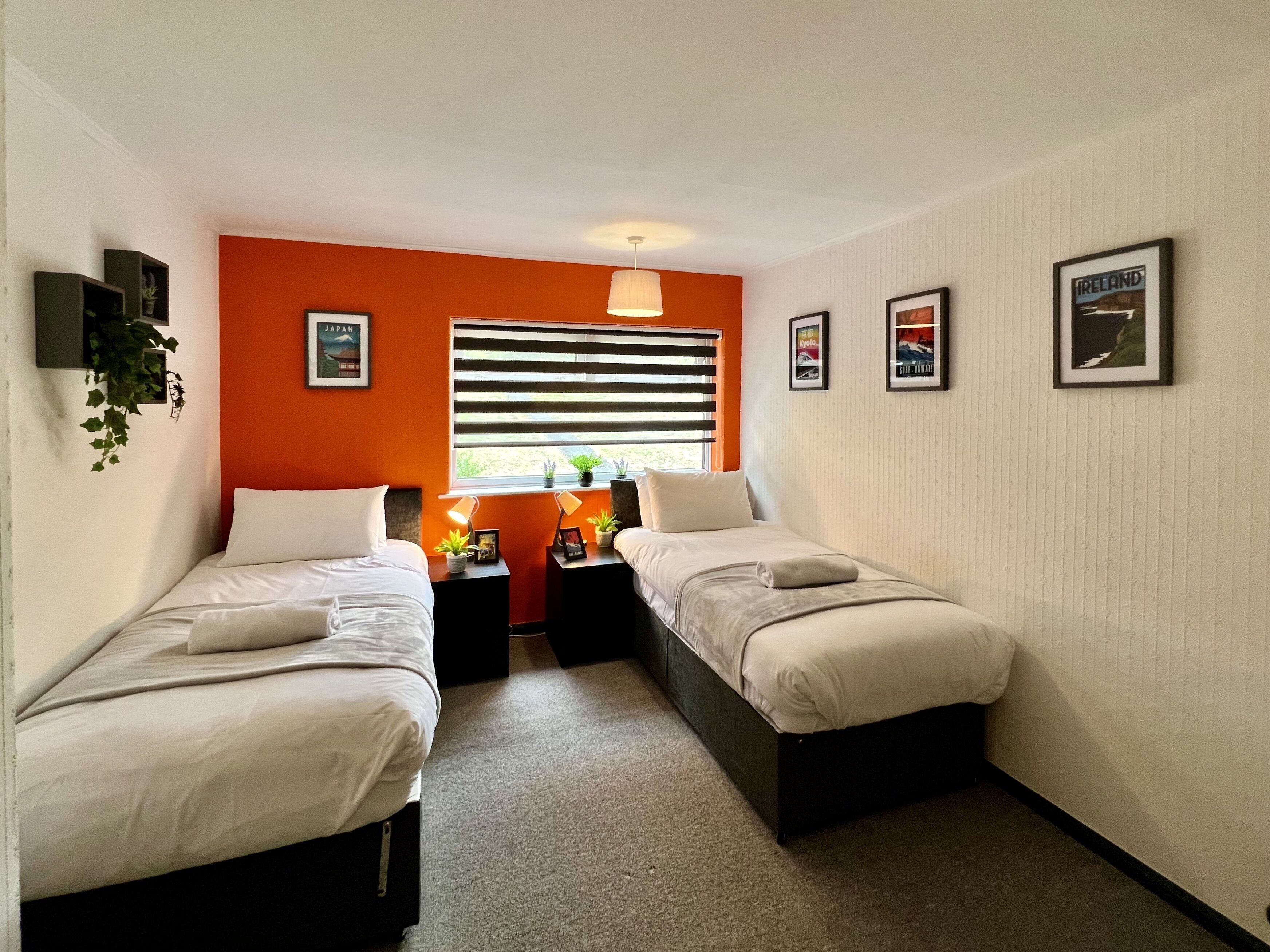 Una habitación con dos camas individuales, una ventana con persianas, una mesita con lámpara y cuadros en la pared.