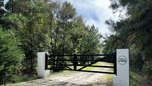 A gated entrance with a brick pillar and a metal gate, surrounded by dense trees and a clear sky. - Escape to Tranquility 125 Acre Property with Trails Lake Spacious House Fire Pit (Kingsland)