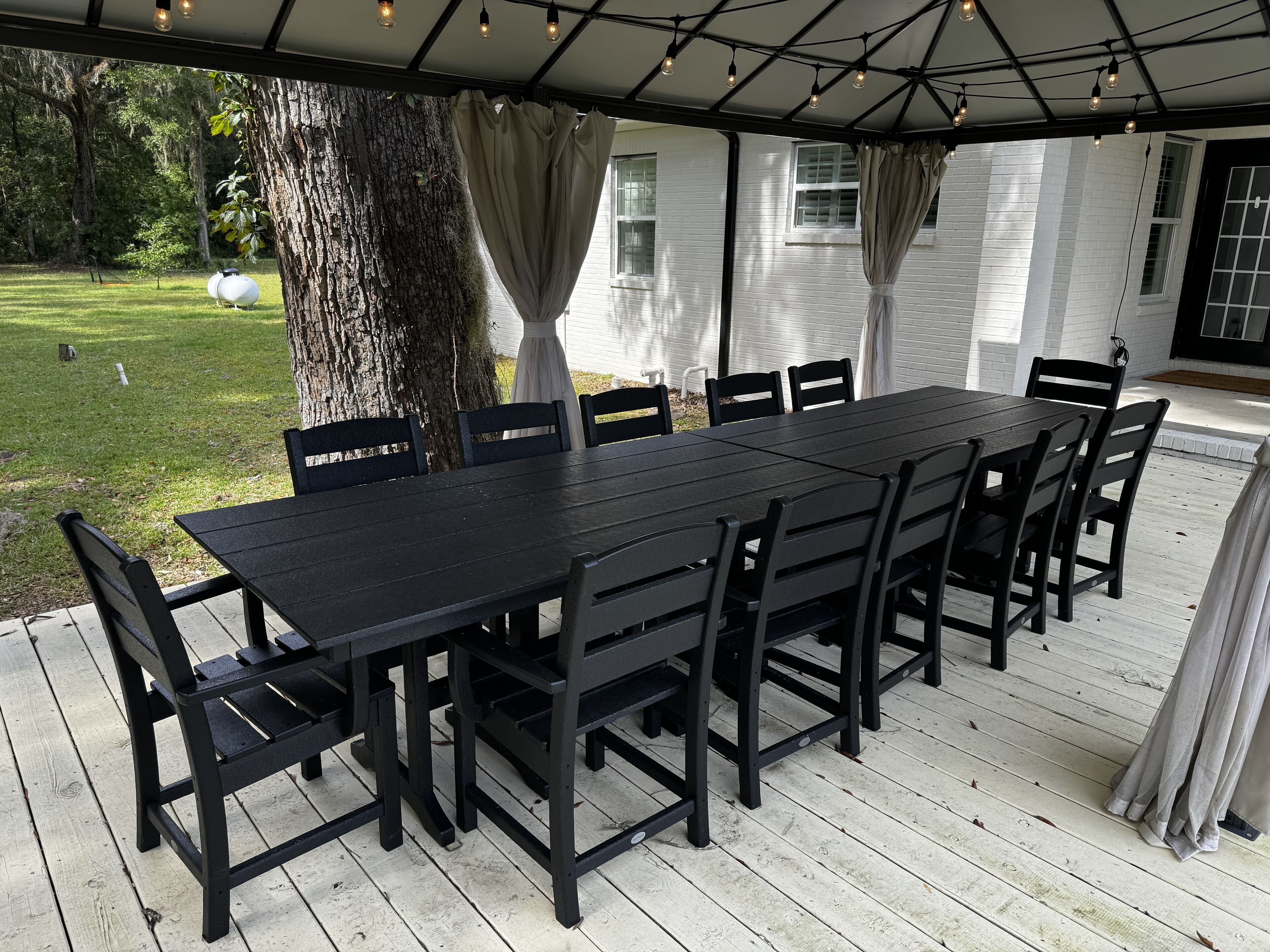 A covered outdoor patio with a black table and chairs, surrounded by white walls and a tree.