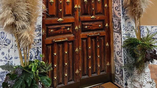 A wooden door with decorative metal studs, flanked by wall-mounted lanterns and potted plants.