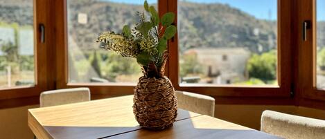 A wooden table with a woven basket vase containing green plants, set against a backdrop of a mountainous landscape through large windows.