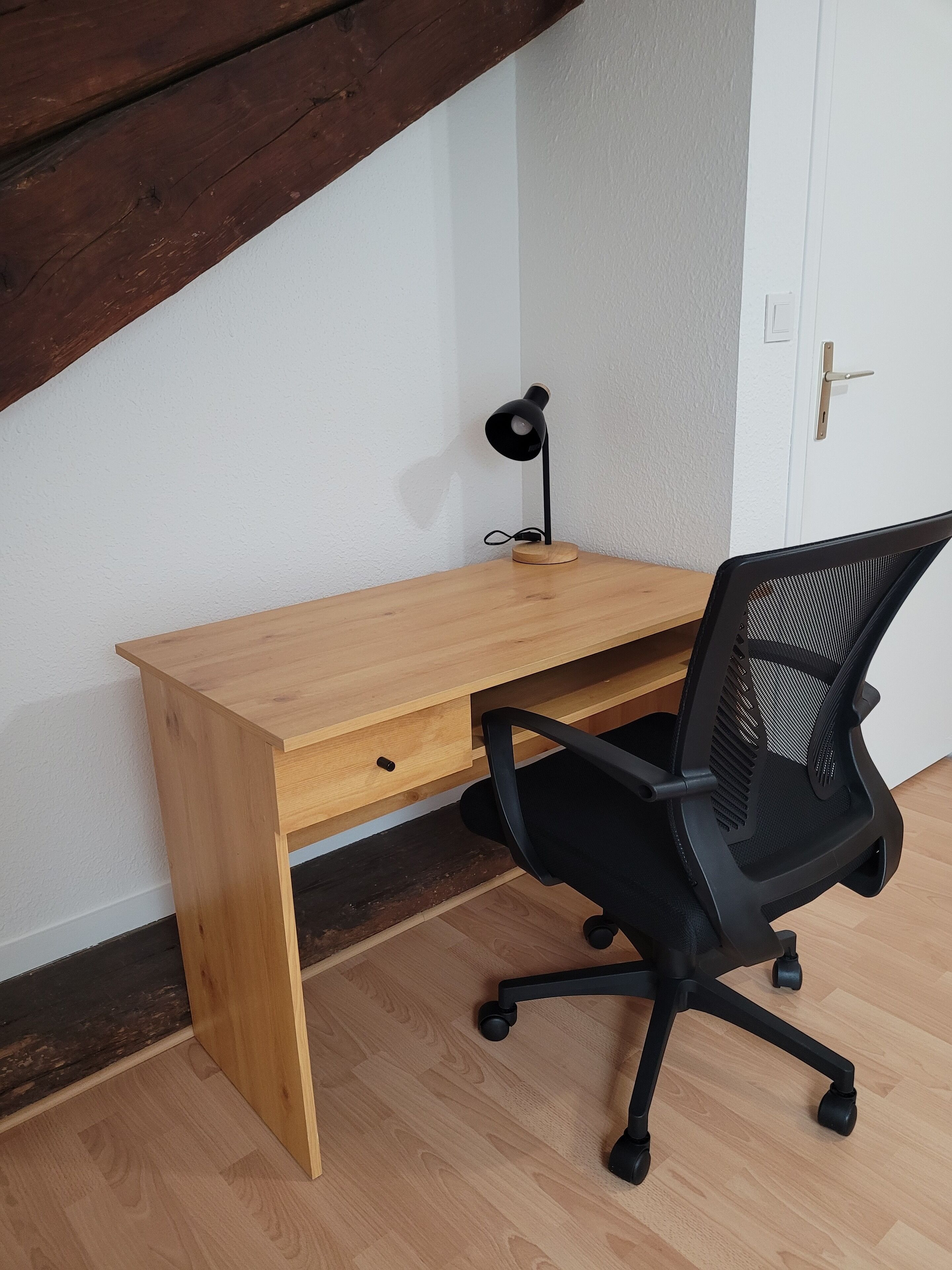 A wooden desk with a black office chair, a black desk lamp, and a white door in a room with wooden flooring and a white wall.