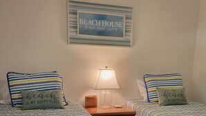 Ein Schlafzimmer mit zwei Betten, jeweils mit gestreifter Bettwäsche und Kissen mit der Aufschrift 'Der Strand ist mein glücklicher Ort'. Ein gerahmtes Schild darüber trägt die Aufschrift 'BEACH HOUSE Meer, Sand, Wellen'.