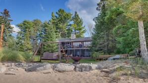 A cabin with a deck, surrounded by trees and a sandy area.