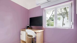 Una habitación con pared morada, un escritorio de madera con una silla, un televisor negro y una ventana que da a árboles y una casa.