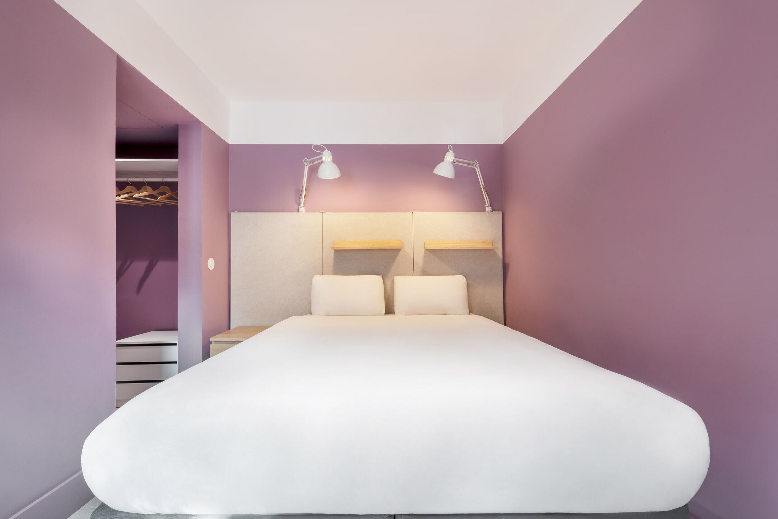 A bedroom with a bed, two bedside lamps, a wardrobe, and a closet with hanging clothes.