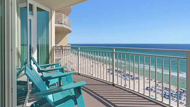 Ein Balkon mit türkisfarbenen Stühlen mit Blick auf einen Strand mit Sonnenschirmen und Menschen.