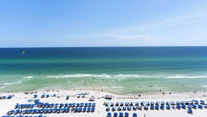 Ein Strand mit Reihen blauer Sonnenschirme und Liegen, einem klaren blauen Himmel und dem Meer im Hintergrund.