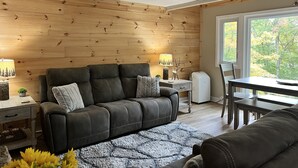 Un salon chaleureux avec un mur en bois, un canapé en cuir foncé, une petite table avec une lampe et une vue sur les arbres par la fenêtre.
