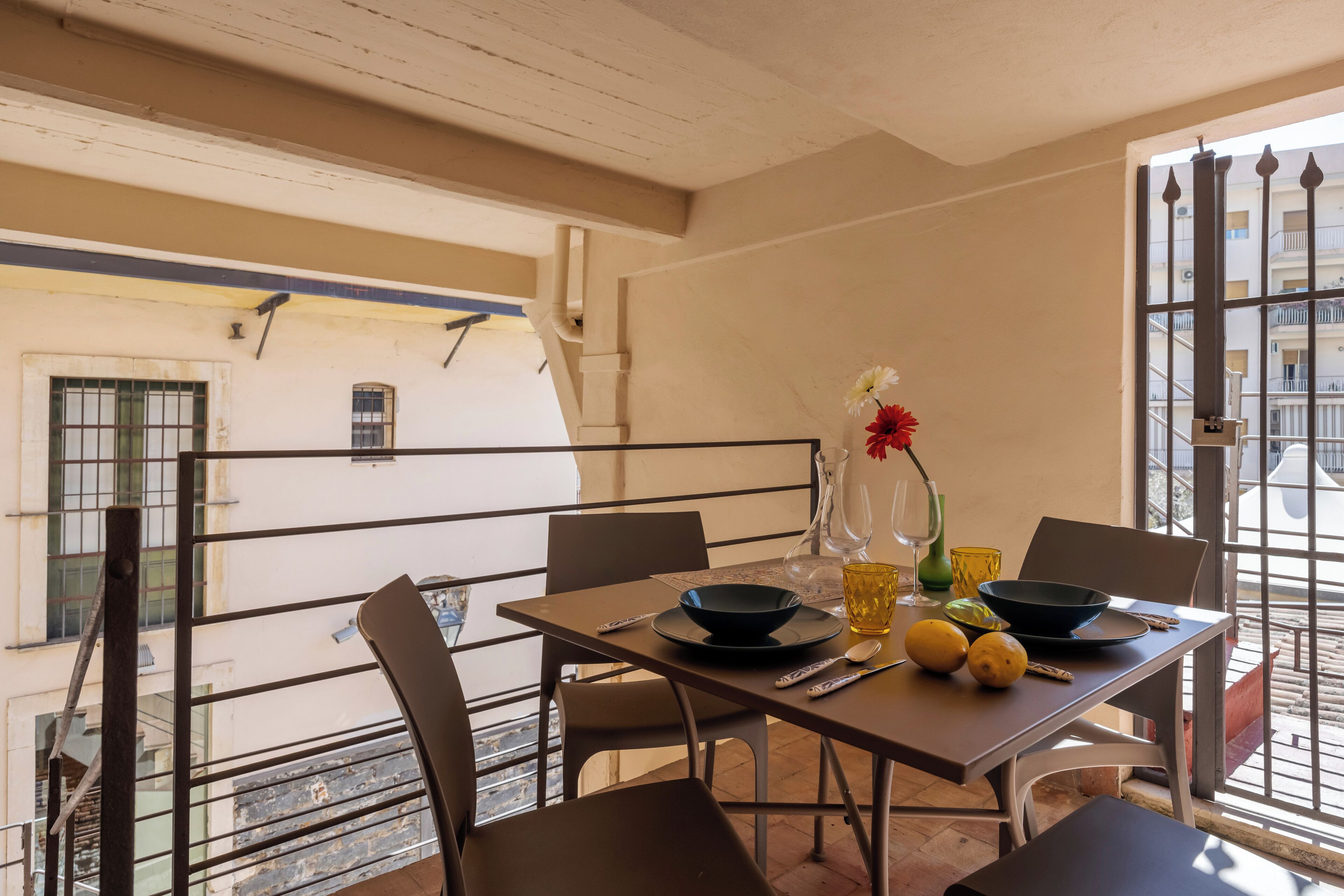 Un coin repas avec une table dressée pour deux personnes, un vase rempli de fleurs et une vue sur la ville, visible à travers une fenêtre ornée de barreaux en métal.