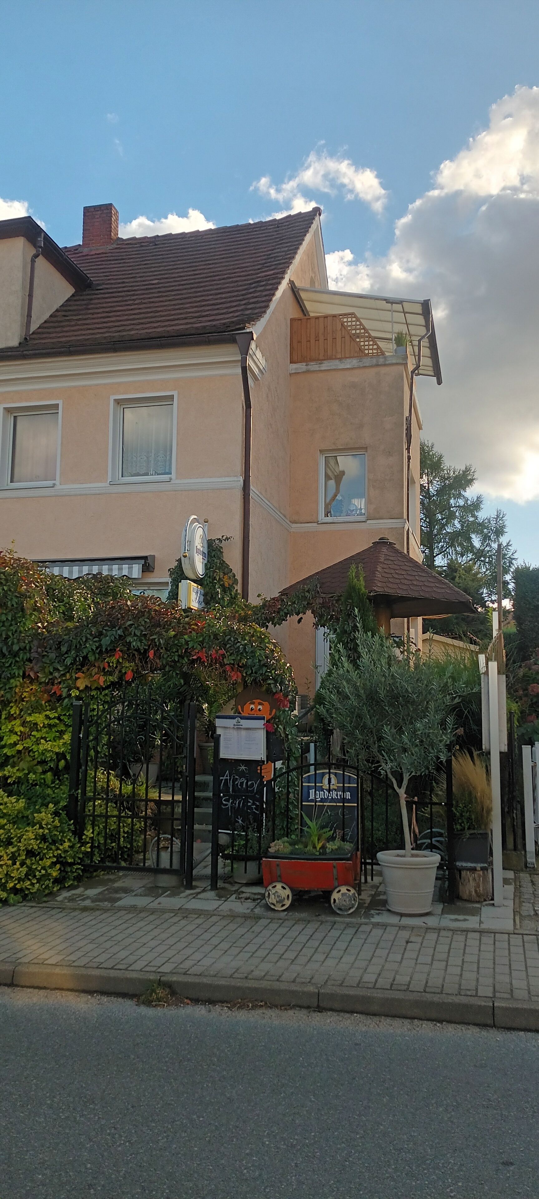Un bâtiment à deux étages, doté d’un toit en tuiles, d’une entrée fermée par un portail et d’un petit jardin agrémenté de plantes en pot et d’un vélo.