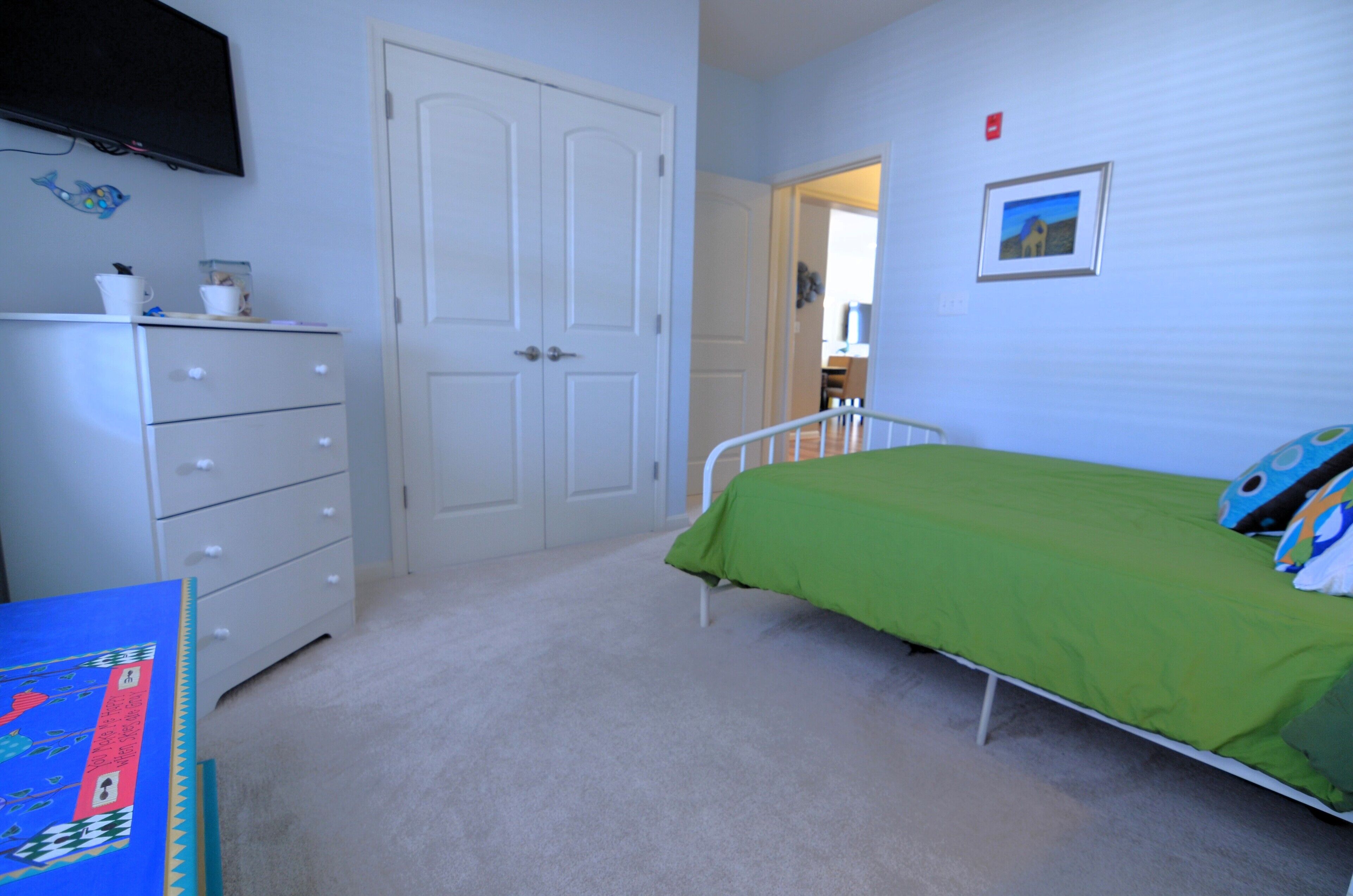 Une chambre avec une couvre-lit verte, une commode blanche et un téléviseur fixé au mur.
