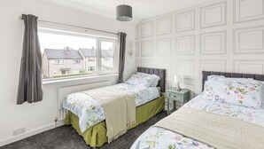 A bedroom with two single beds, a window with curtains, a radiator, and a wall with a geometric pattern. - 3 Bed House - Sleeps 5 - Parking - Garden - Wifi (Blackburn)