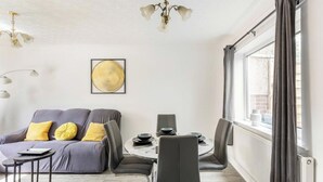 A modern living room with a grey sofa, yellow cushions, a dining table with chairs, and a large window with curtains. - 3 Bed House - Sleeps 5 - Parking - Garden - Wifi (Blackburn)