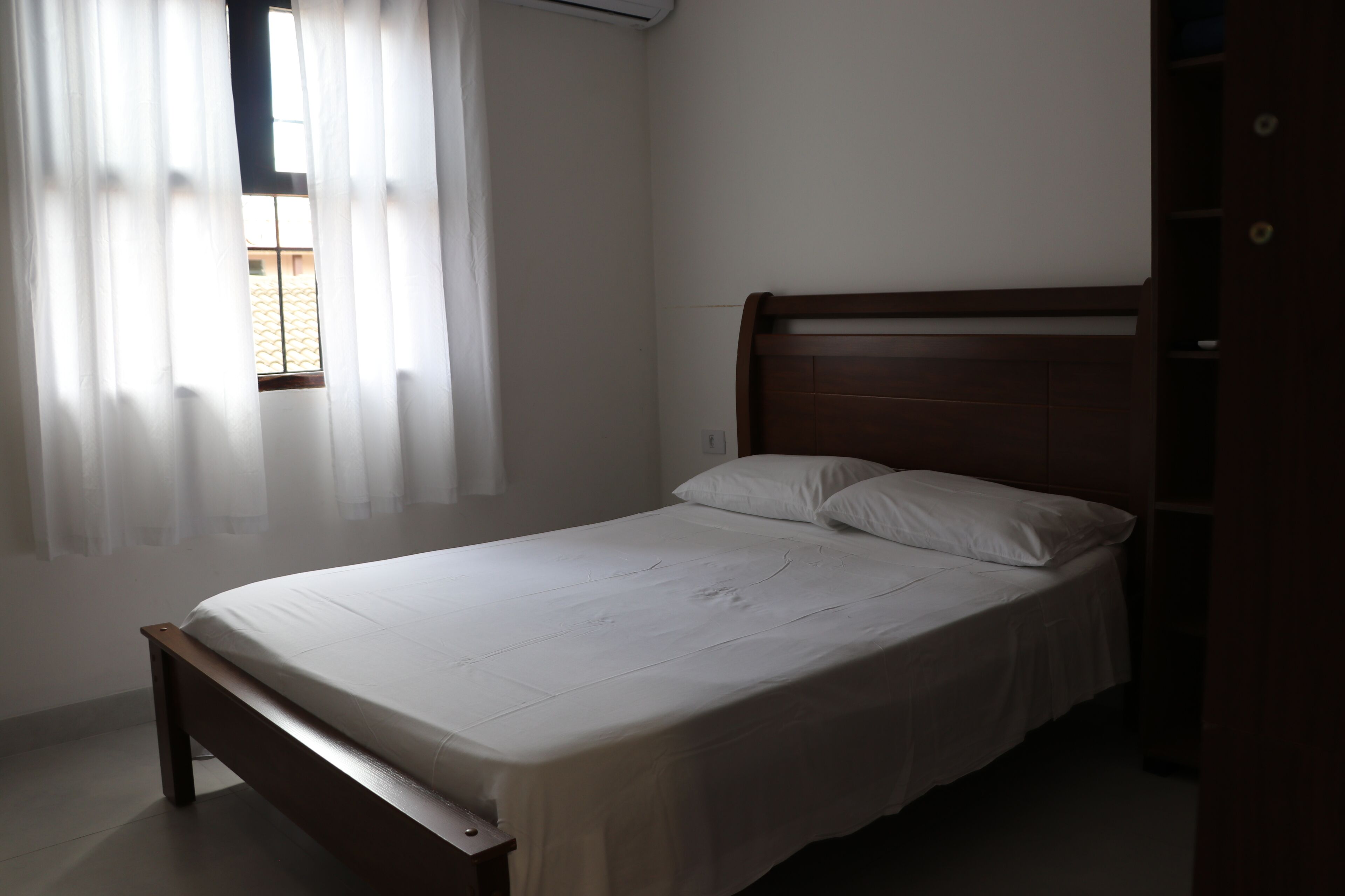 A single bed with a wooden frame and white bedding in a room with white walls and a window with sheer curtains.