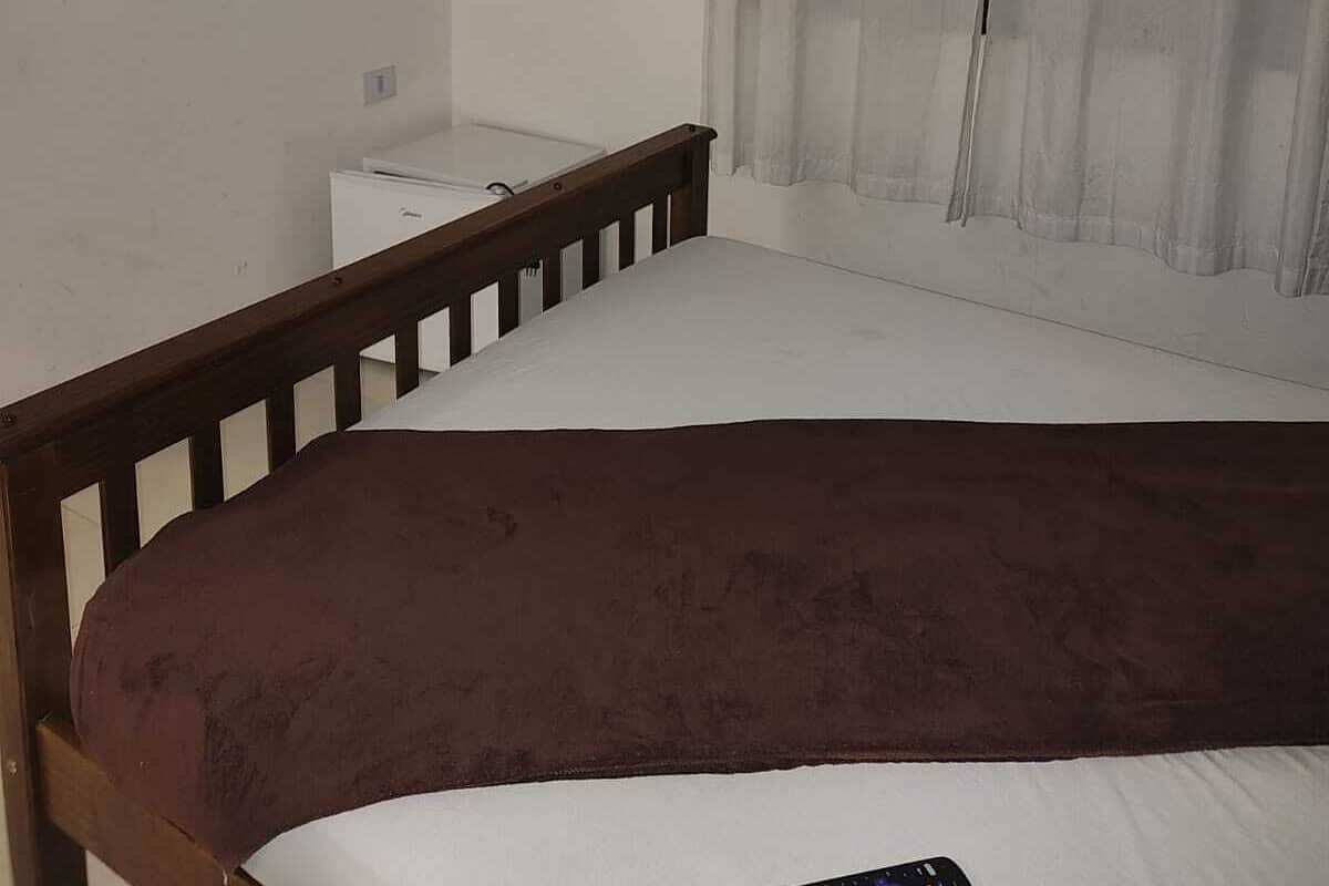 A bedroom with a single bed, a small bedside table, a TV mounted on the wall, and a window with sheer curtains.