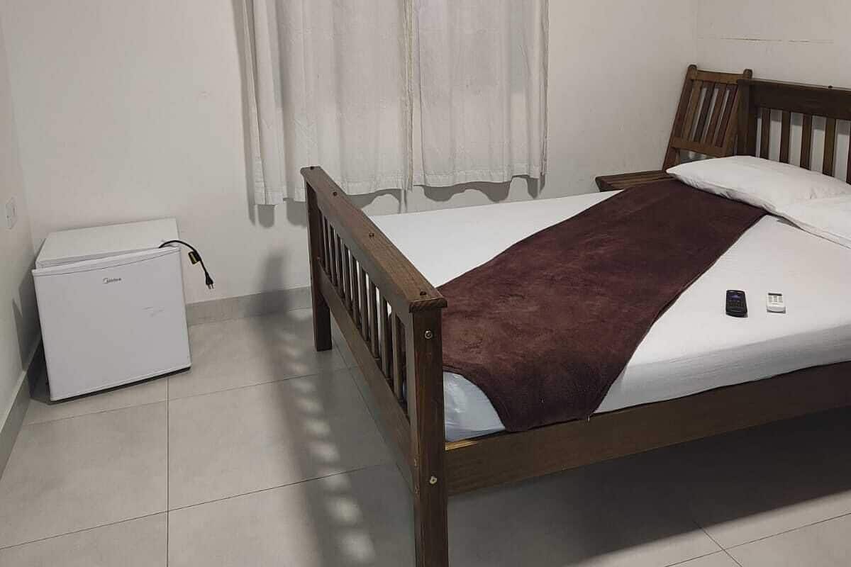 A bedroom with a wooden bed, a white bedside table, a small white appliance, and a window with curtains.