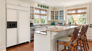 A kitchen with white cabinetry, a central island with bar stools, and a window with a view of trees. - Gated Seaside Community sleeps 10, private courtyard pool & community pool (St Simons Island)