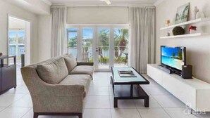 A modern living room with a sofa, coffee table, TV, and a balcony with curtains. - Waterfront Sanctuary Cove, 2Bed 2Bath, King Bed, Pool, Porch at Grand Harbour. (Grand Harbour)