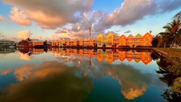 A row of colorful buildings reflecting on calm water, with a clear sky above. - Waterfront Sanctuary Cove, 2Bed 2Bath, King Bed, Pool, Porch at Grand Harbour. (Grand Harbour)