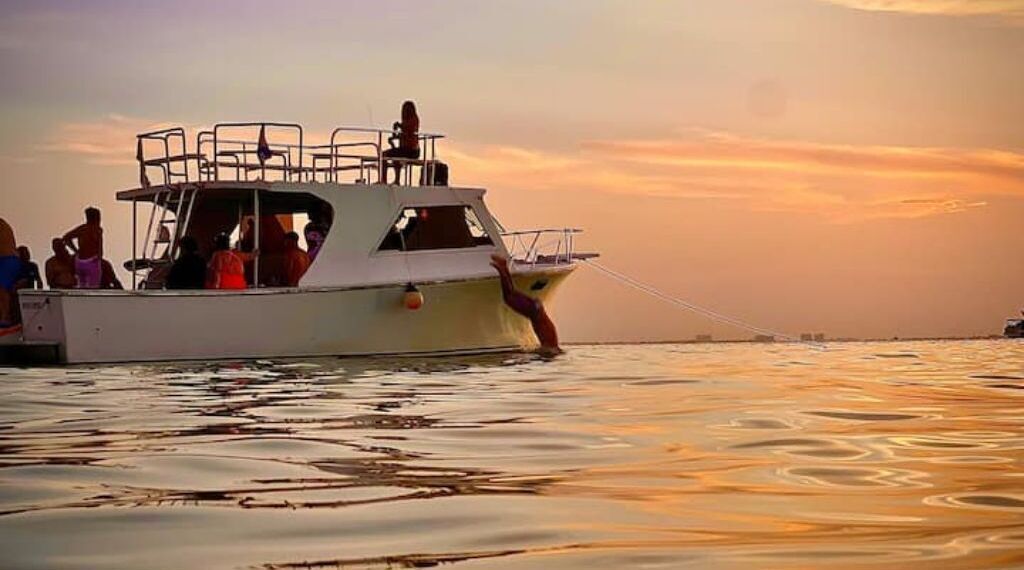 A boat with people on board, a person leaning out of the side, and a sunset in the background.