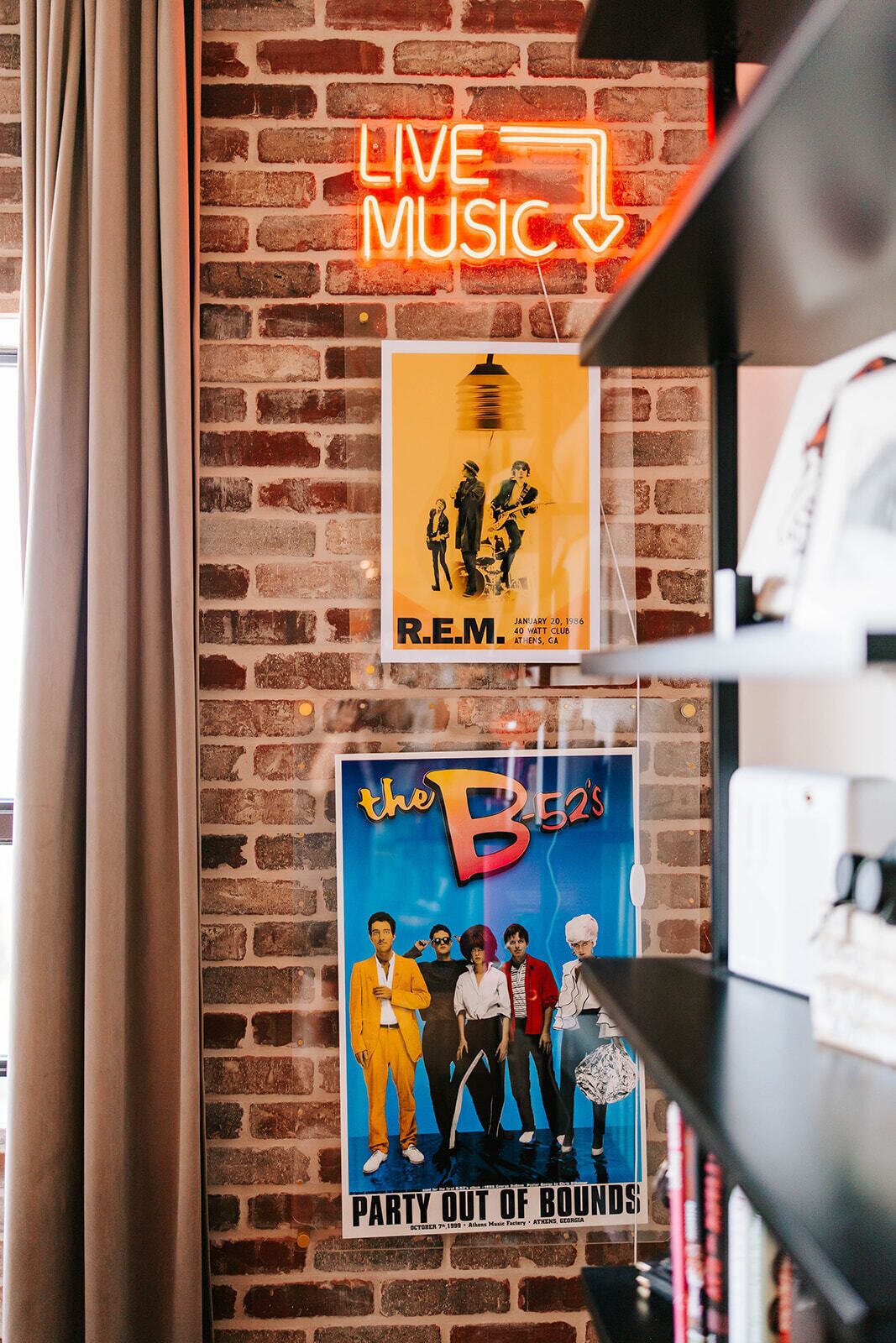 A brick wall with a neon sign reading 'LIVE MUSIC' and two posters, one for R.E.M. and another for The B-52's.