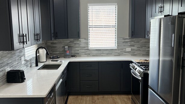 Una cocina moderna con armarios oscuros, un refrigerador de acero inoxidable, un horno empotrado y una ventana con persianas.