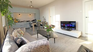 Un salon moderne avec un canapé, une table basse et un téléviseur. On aperçoit une cuisine au fond, avec un coin repas.