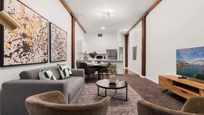 A modern living room with a grey sofa, two armchairs, a coffee table, and a TV on a wooden stand. There are abstract paintings on the wall and a cityscape on the TV screen. - Heart of Darling Harbour (Pyrmont)