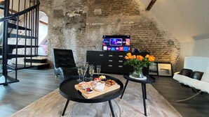 A loft-style living room with exposed brick walls, a black leather sofa, a round black coffee table with snacks, and a flat-screen TV mounted on the wall. - Spacious cosy apartment in historic building in the centre of charming Vianen (Vianen)