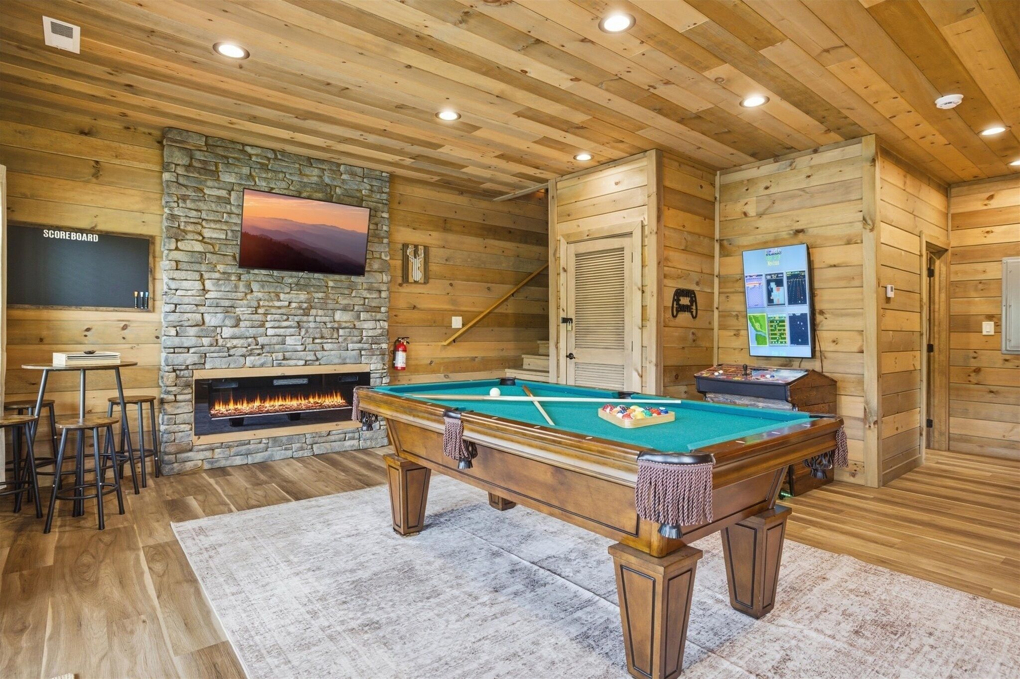 A pool table with a fire feature, a TV mounted on a stone wall, and a bar area with stools.