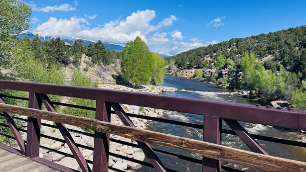 Un pont en bois, doté d’une rambarde métallique, surplombe une rivière, avec des montagnes en toile de fond.