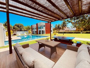 A covered outdoor seating area with a pool, wicker furniture, and a view of a modern building. - Casa Tatuamunha Beach (Porto de Pedras)