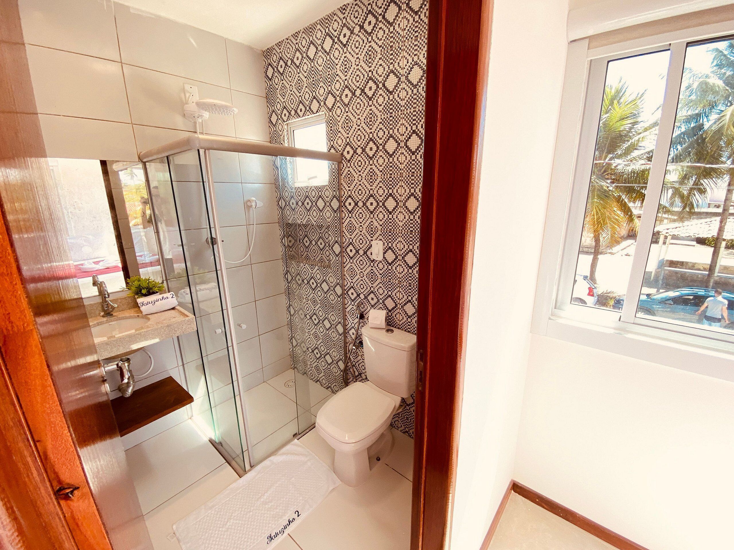 A bathroom with a glass shower enclosure, a toilet, and a window with a view of palm trees.