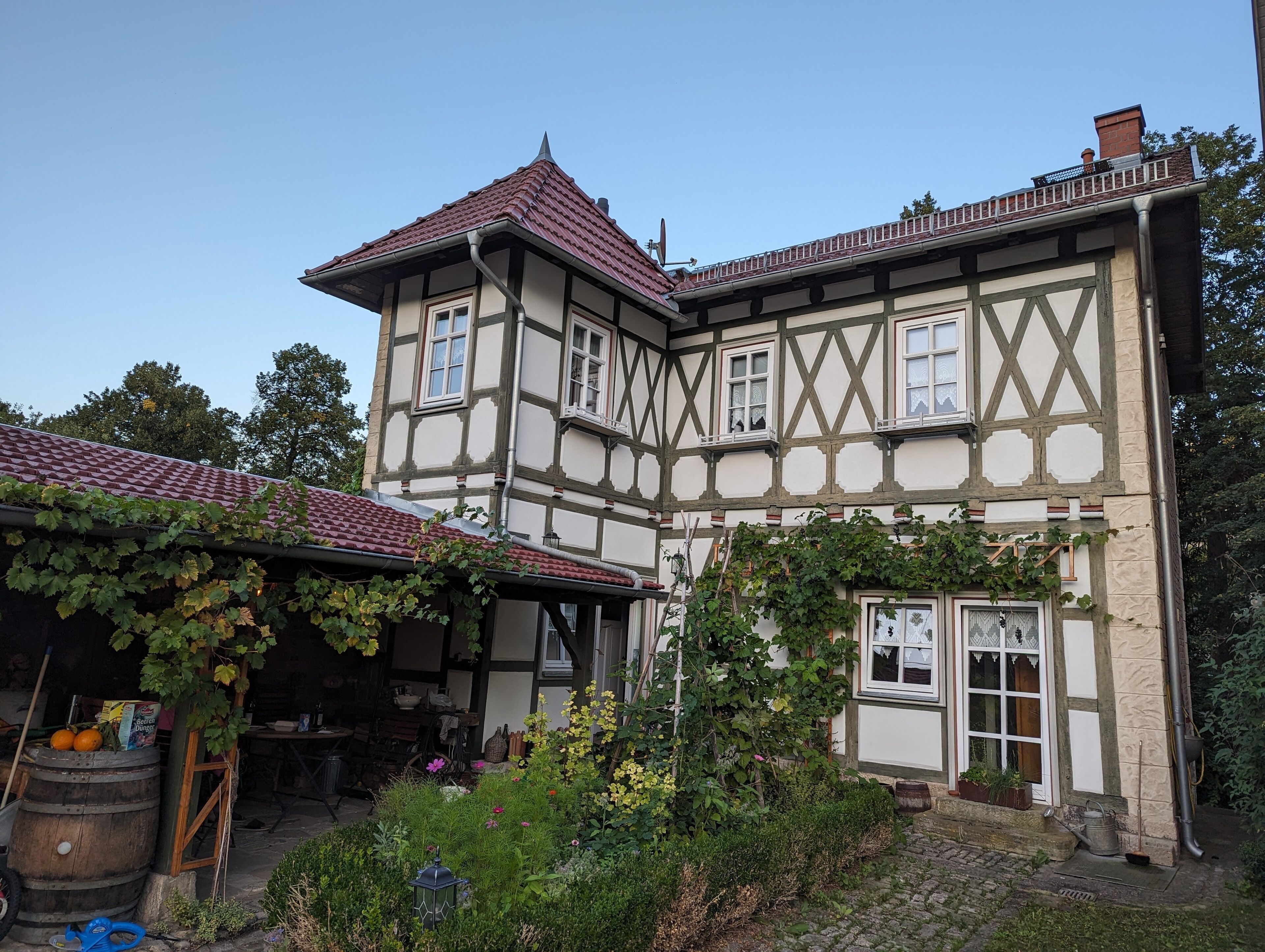 Ein traditionelles Fachwerkhäuschen mit geneigtem Dach, einem Garten mit verschiedenen Pflanzen und einem Fass.