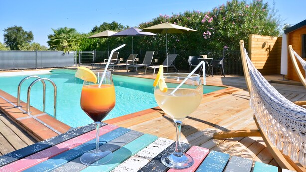 A poolside area with two drinks on a table, lounge chairs, and umbrellas.