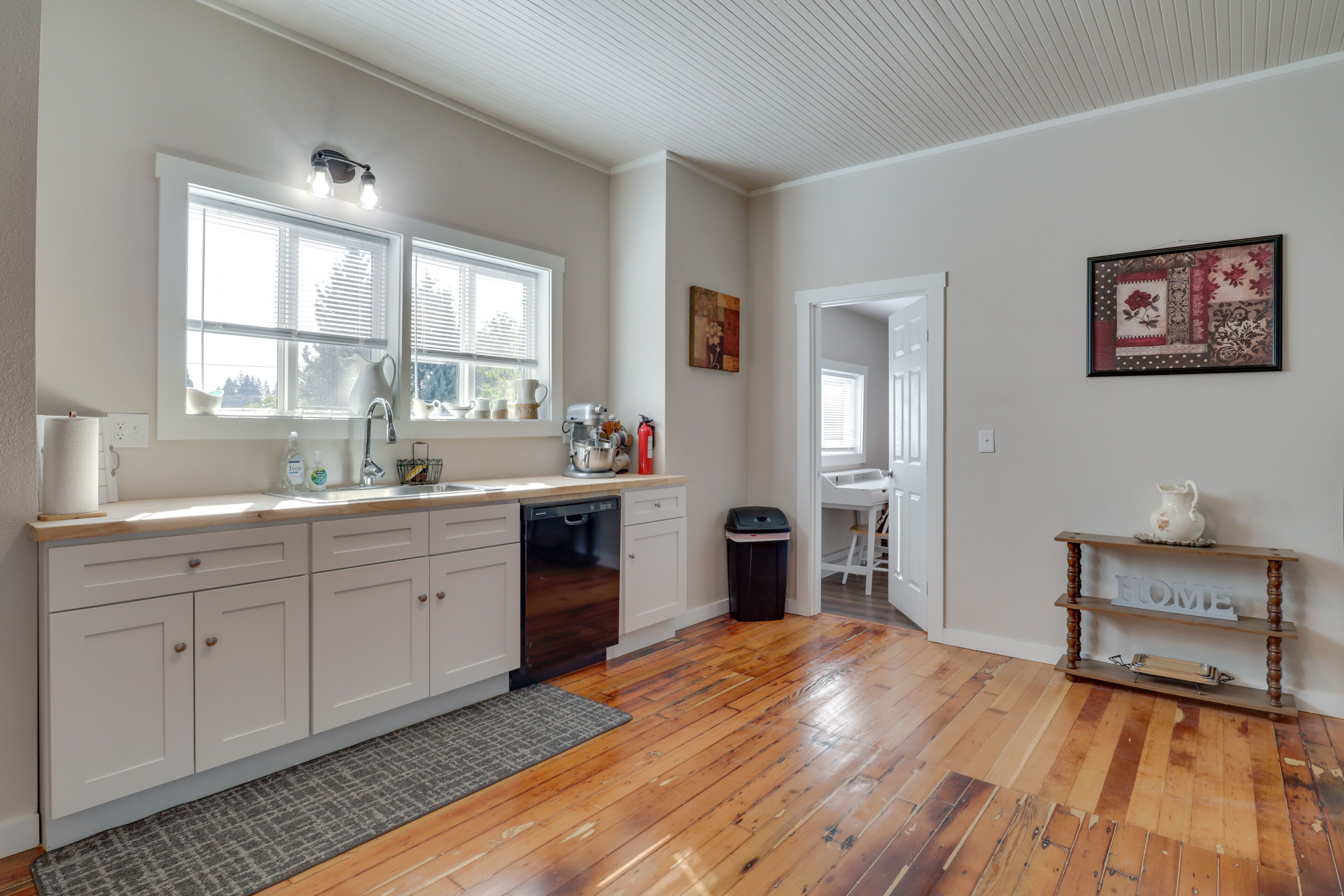 A spacious kitchen with wooden flooring, white cabinets, and a large window with blinds.