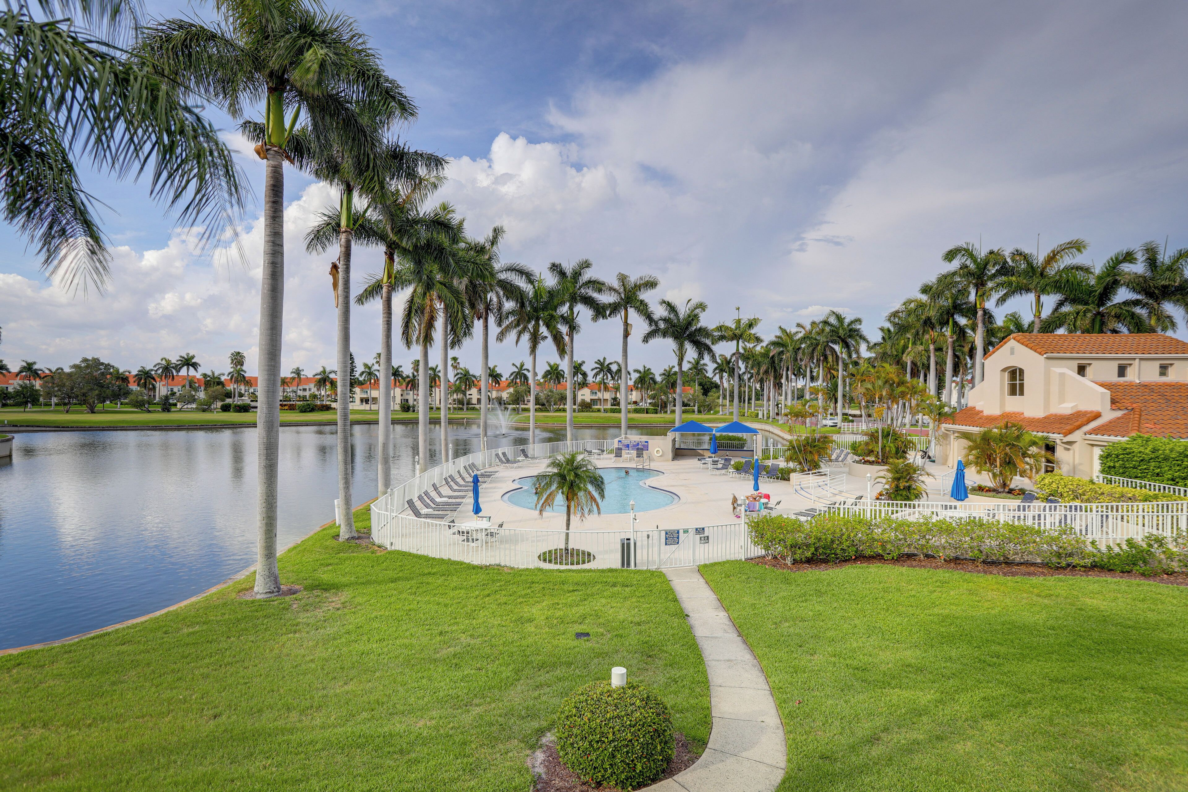 A pool area with palm trees, a water body, and a building with a red roof.