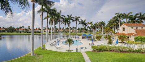 A pool area with palm trees, a water body, and a building with a red roof.