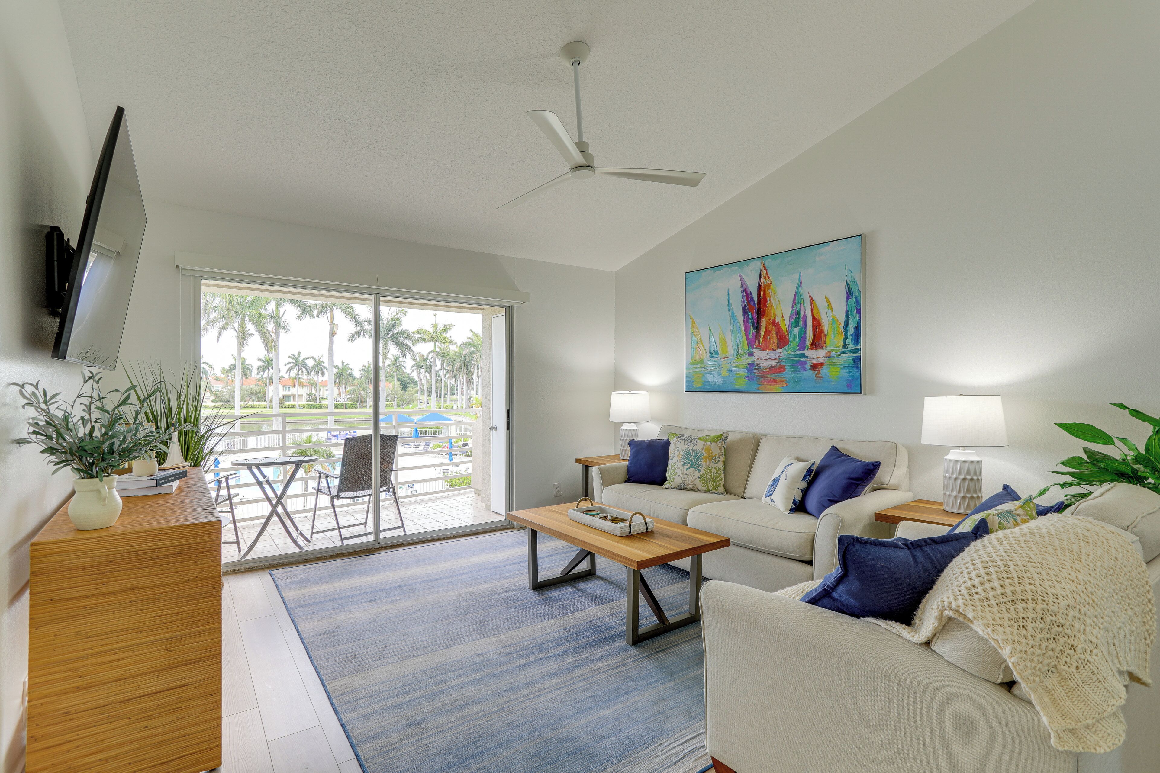 A living room with a sofa, coffee table, and a painting of sailboats. There is a TV mounted on the wall and a ceiling fan.