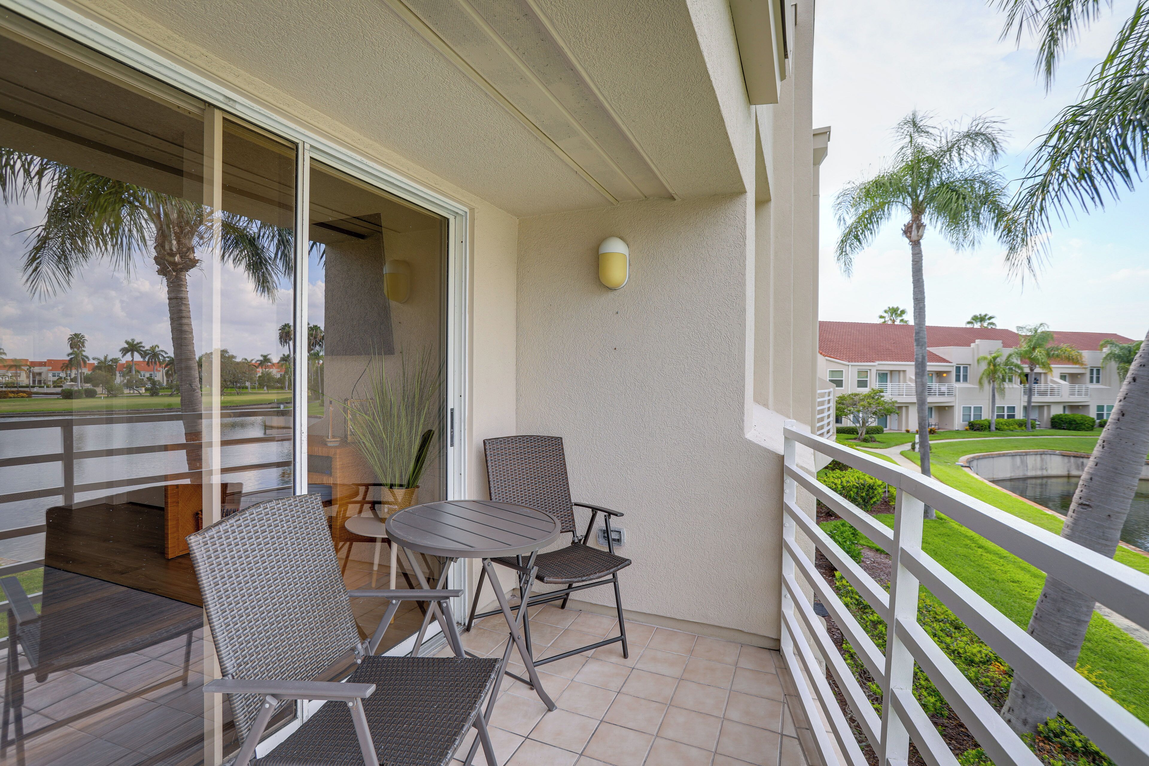 A balcony with a table and chairs, a potted plant, and a view of a landscaped area with palm trees and a building in the background.