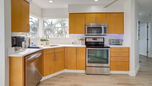 A modern kitchen with wooden cabinets, stainless steel appliances, and a large window offering a view of palm trees. - Sunny St Pete Getaway With Shared Pool & Hot Tub (St. Petersburg)