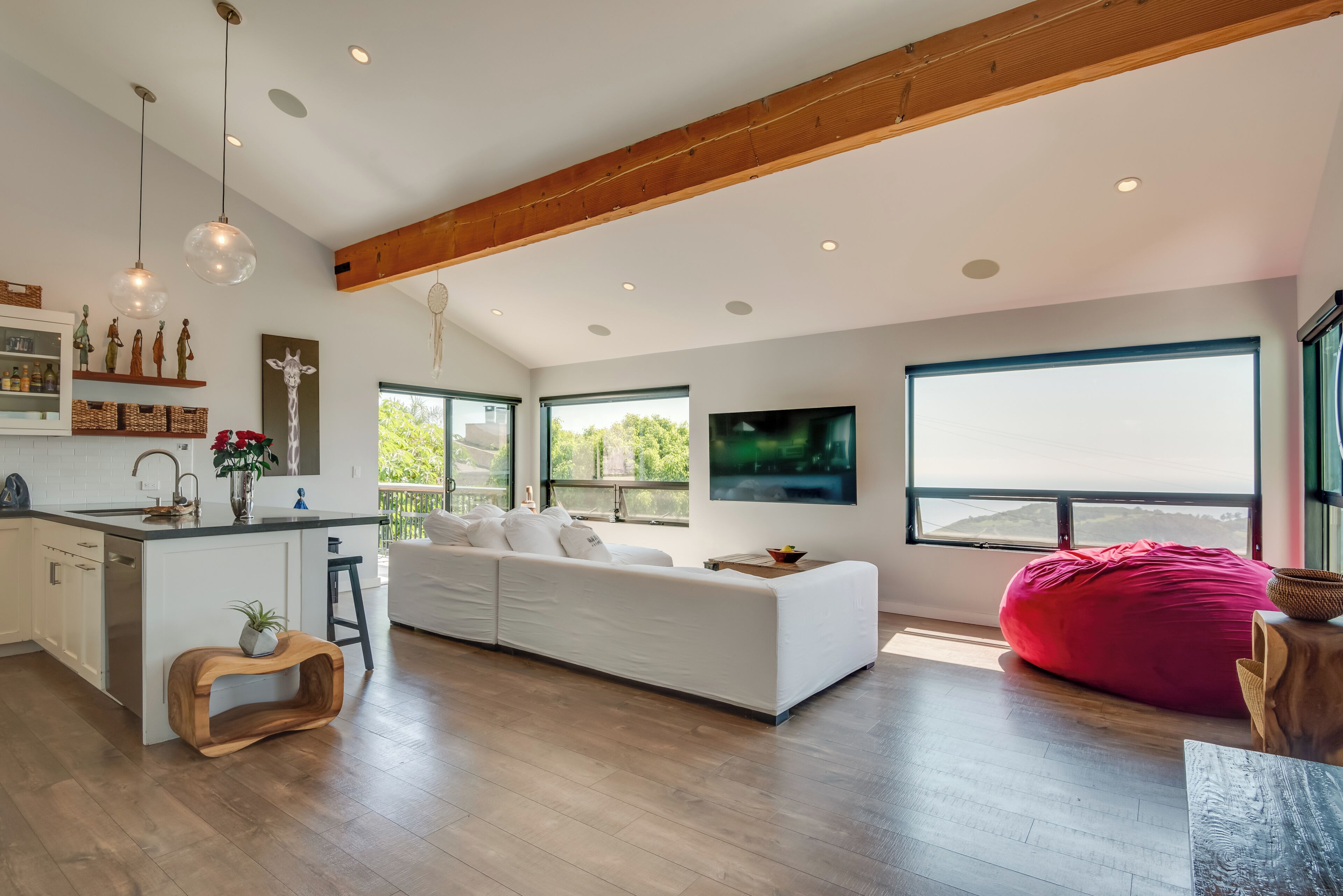 A modern living room with a white sofa, a red bean bag, and a kitchen area with wooden beams and pendant lights.