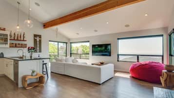A modern living room with a white sofa, a red bean bag, and a kitchen area with wooden beams and pendant lights.