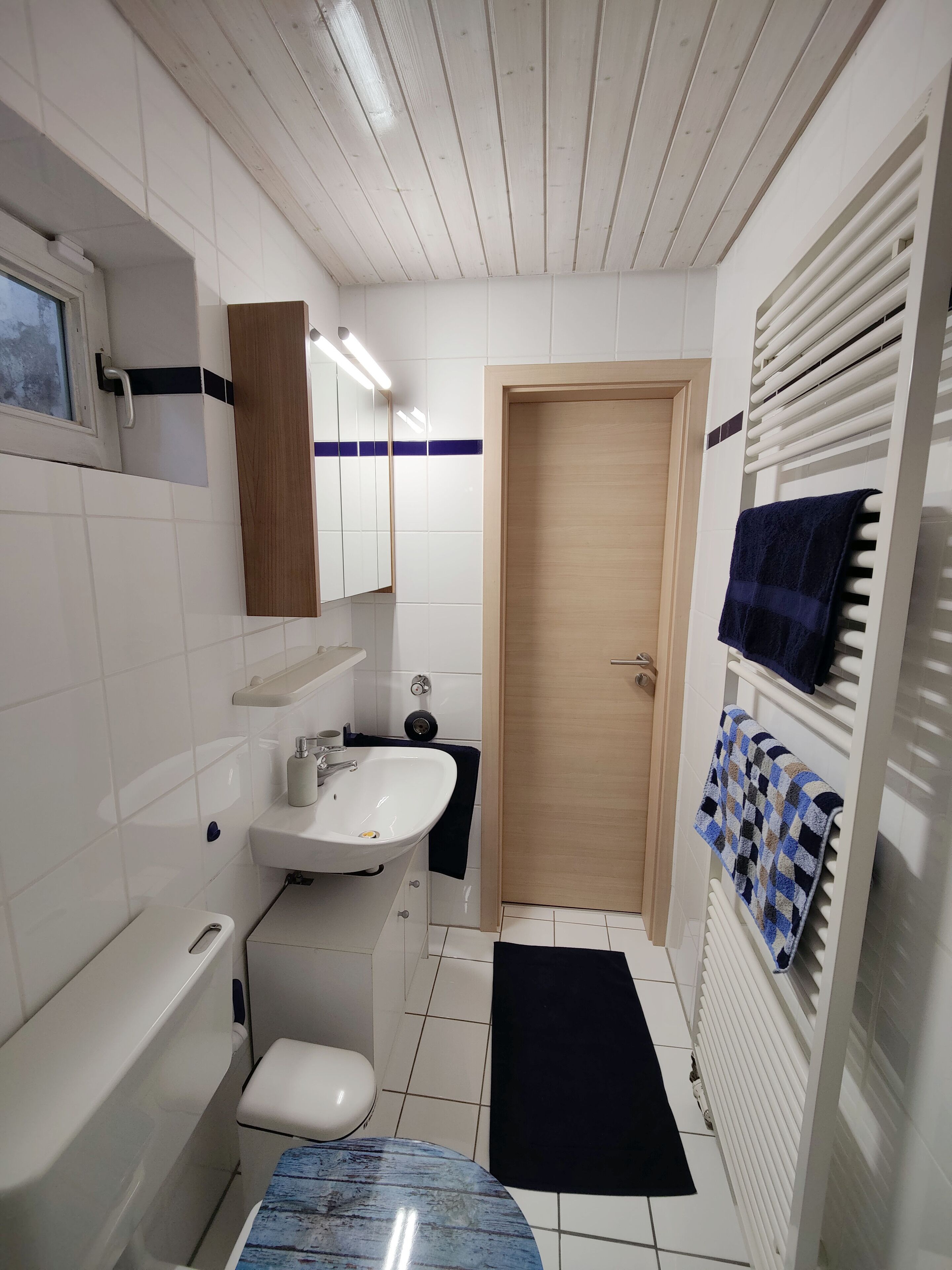 Une petite salle de bain propre, avec des carreaux blancs, une porte en bois, un lavabo blanc, des toilettes et un porte-serviettes avec des serviettes.