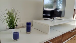 Un salon moderne avec un téléviseur à écran plat posé sur une console blanche, une plante en pot et un porte-bougie bleu.