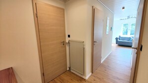 Un couloir avec un plancher en bois, des murs blancs et un radiateur. Il y a une porte sur la gauche et une fenêtre au fond.