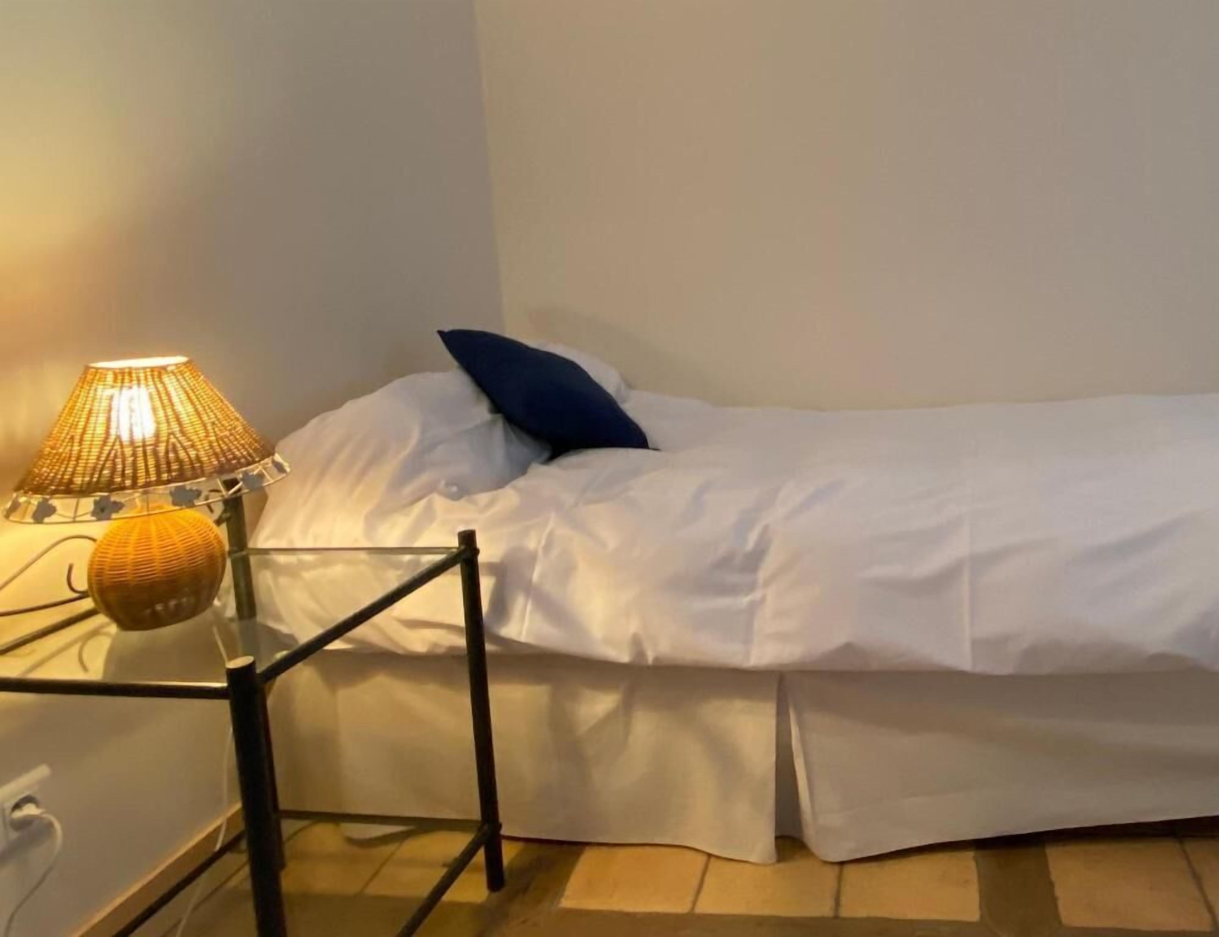 A single bed with white bedding and a blue pillow, a bedside lamp with a woven shade, and a small round object on the nightstand.