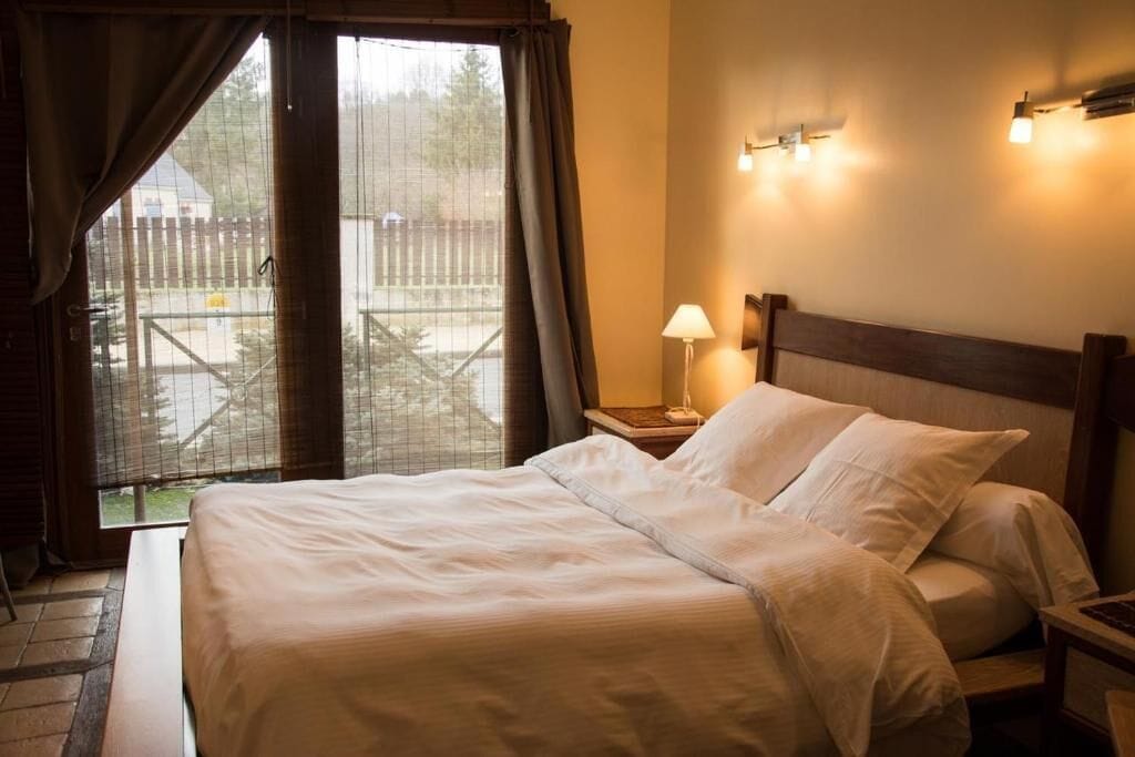 Habitación de hotel con cama, mesita de noche, lámpara y vista a través de una ventana con cortinas.