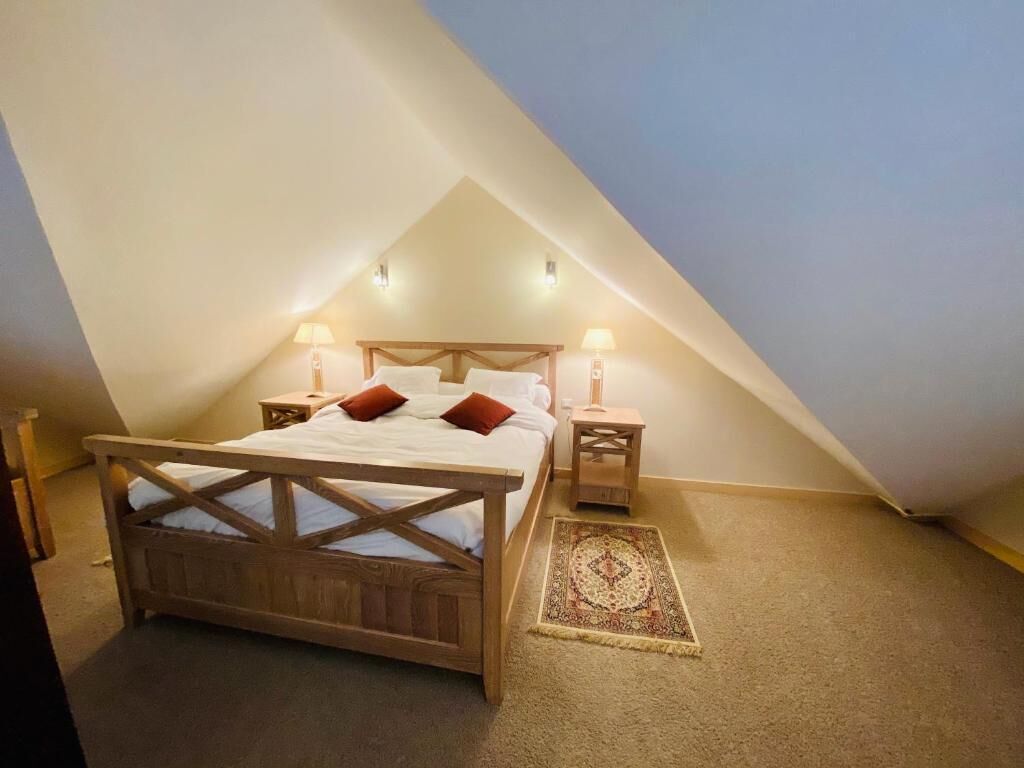 Een slaapkamer met een schuine plafond, een houten bedframe, twee bedlampen en een kamerbreed tapijt met dessin.